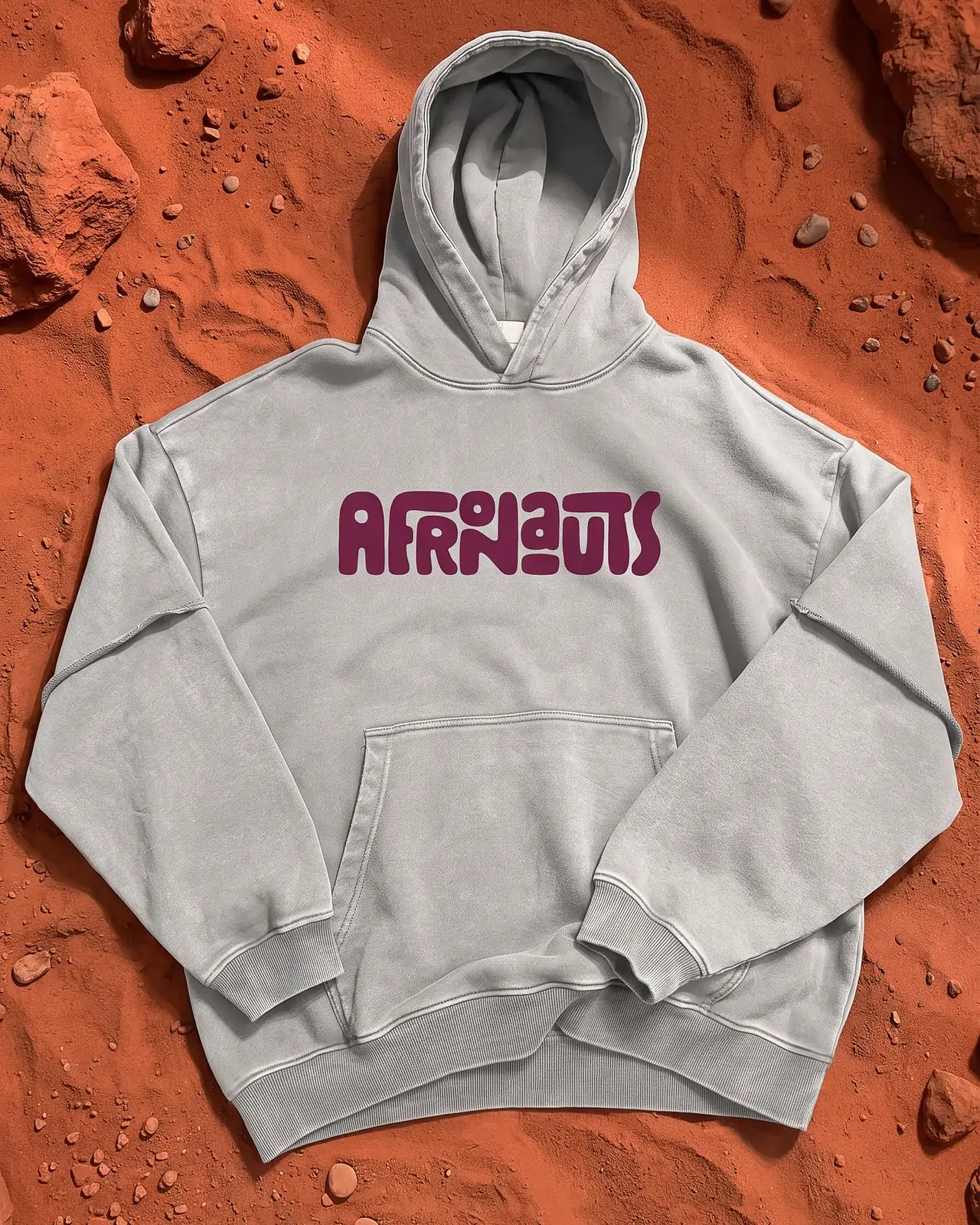 A light grey Afronauts hoodie laid flat on red, rocky ground styled as the surface of Mars.
