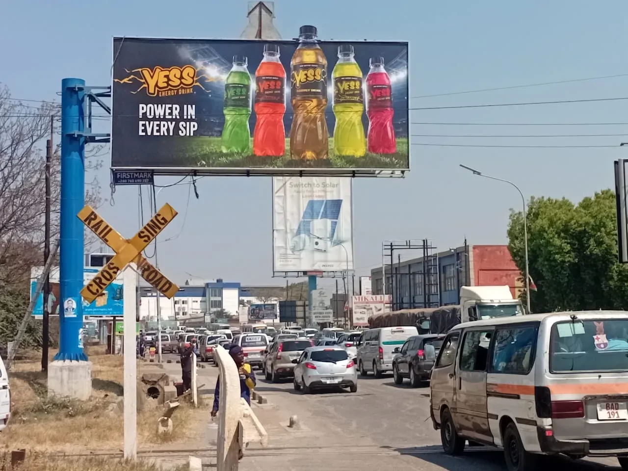 A YESS billboard above traffic beside a railway crossing sign.